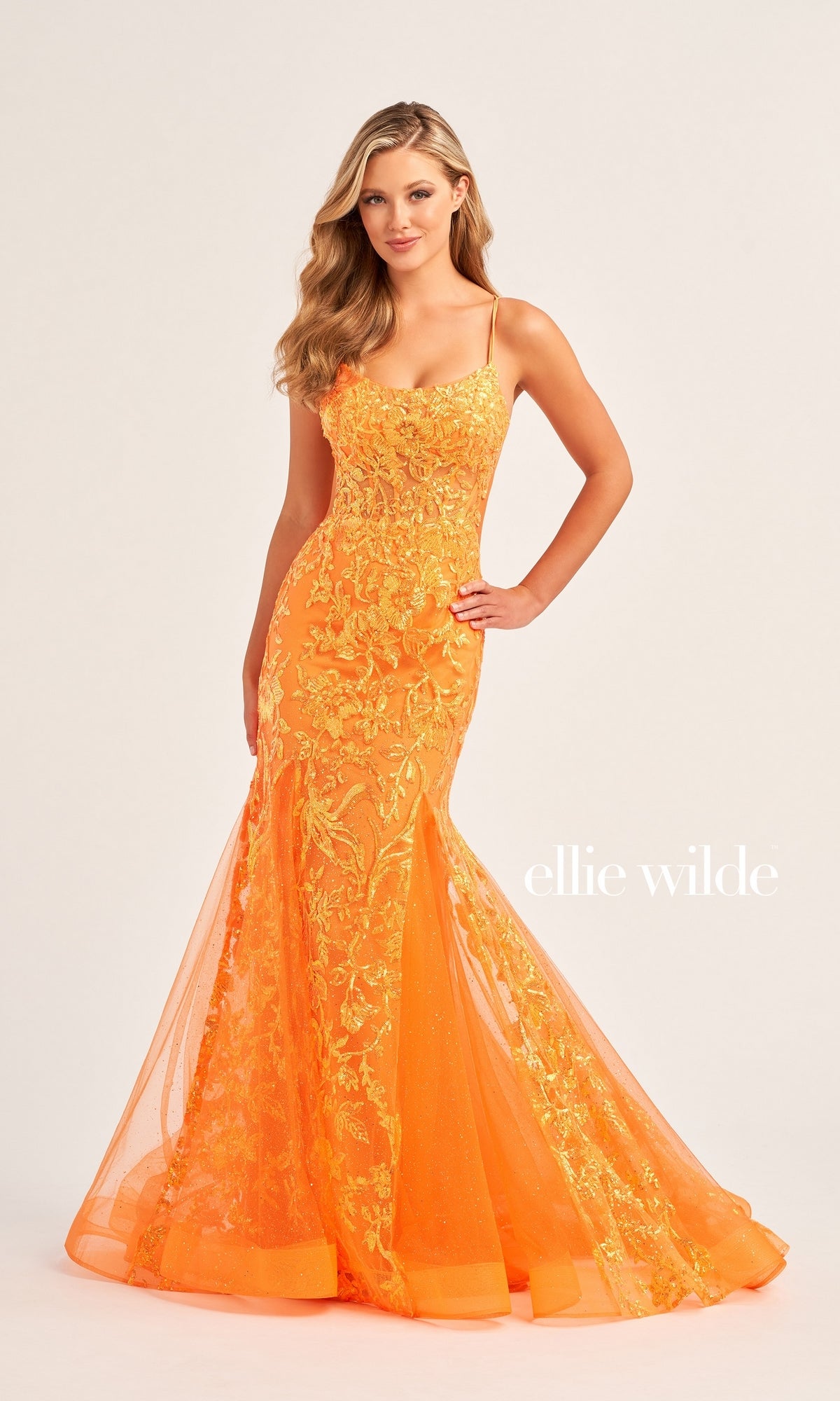 A woman wears the fitted Ellie Wilde Sequin Long Mermaid Prom Dress EW35008, a bright orange mermaid gown with floral embroidery, standing against a plain background.